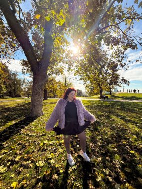 Sonbahar ağaçlarının altında gün ışığıyla gülümseyen bir kadın. Neşe, açıklık ve doğayla uyum içinde kendini ifade etme.
