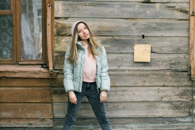 Nane rengi ceketli ve pantalonlu bir kadın ahşap bir evin penceresinde poz veriyor. Basitlik, gençlik ve klasik cazibeyle sakin modern tarz.