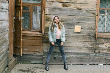 Nane rengi ceketli ve pembe kapüşonlu bir kadın eski ahşap evin karşısında duruyor. Sakin bir ifade, şehir melankolisi ve minimalist kış havası.