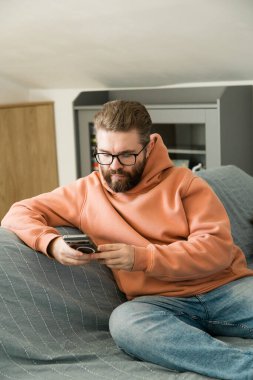 Kanepede oturan ve akıllı telefon kullanan bir adam. Ev ortamında dijital rahatlama, iletişim, bilgi taraması ve boş zaman aktivitesini temsil eden modern yaşam tarzı sahnesi