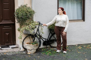 Bitkilerle süslenmiş bisikletle duvarın yanında duran bir kadın. Şehir yaşamı, sonbahar sükuneti ve yavaş şehir ritmi.