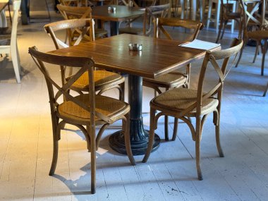 Pencere kenarında güneş ışığında sandalyeli ahşap kafe masası. Sessiz ortam, tasarım dengesi ve günlük konukseverlik ritmi.