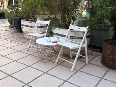İki beyaz sandalye ve fayanslı sokak terasında yuvarlak masa. Basitlik, sessiz rahatlama ve açık kafe hayatının günlük cazibesi.