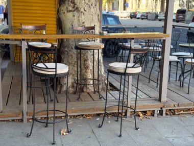 Açık bar tabureleri ve ağaç gölgesinin altında yüksek masalar. Açık teras ortamında şehir eğlencesi, mimari ve günlük yaşam tarzı