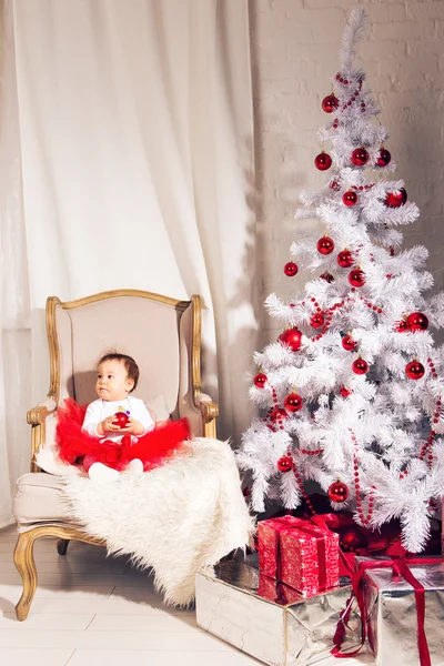 Menina senta-se em uma cadeira perto da árvore de Natal — Fotografia de Stock