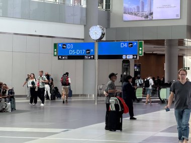 Havaalanı terminalinde yön işaretleri ve merkezi saat altında yürüyen yolcular. Zaman yönetimi, yol bulma netliği, seyahat koordinasyonu ve ulaşım merkezinde organize yolcu akışı..