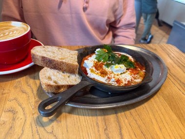 Shakshuka ile kahvaltı, ahşap masada ekmek ve kapuçino. Konforlu kafe ortamında yiyecek ve Akdeniz aromaları
