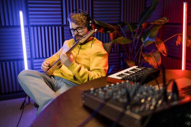 Odaklanmış bir erkek müzisyen, çağdaş bir kayıt stüdyosundaki dijital ses düzenleme kurulumunun arka planına karşı bir bas gitar ayarlar..