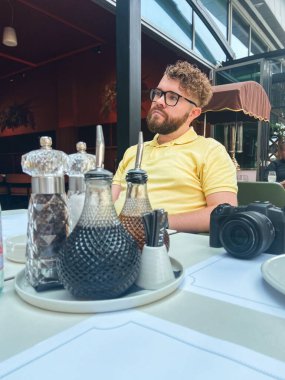 Kafe masasında oturan adam ön planda kamera ve sofra takımı ile. Şehir eğlencesi, yaratıcı yaşam tarzı, sokak fotoğrafçılığı ve rahat sosyal atmosfer..