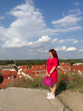 Mavi gökyüzünün altında kırmızı çatılara bakan pembe çantalı bir kadın. Seyahat bakış açısı, şehir keşfi anı ve kişisel yaşam tarzı yansıması.