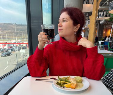 Pencerenin yanındaki kafede yemek yiyip şarap içen bir kadın. Dengeli beslenme, sakin yemek, düşünceli yaşam tarzı, şehir manzarası, ve kapalı alandaki günlük zevkin huzurlu anı