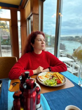 Kahve içerken pencerenin kenarında oturan bir kadın kahvaltıda dışarı bakıyor. Sessiz bir yansıma, sakin bir sabah, duygusal denge ve şehrin kıyısı yakınında huzurlu bir yaşam tarzı anı.