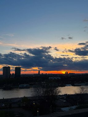 Gün batımında gökyüzü dramatik bulutların altındaki bir nehri yansıtıyor. Şehir manzarası, akşam sakinliği, zaman geçişi, atmosferik ışık, mimari ve doğa arasındaki denge ve sessiz yansıtıcı anlar.