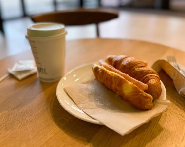 Sosisli kruvasan sandviç ve sehpanın üzerinde kahve. Basit kahvaltı, gündelik yemek, sıcak atmosfer ve günlük yemek rutini.