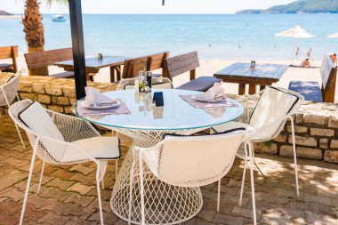 Beyaz sandalyeli ve cam yüzeyi olan deniz kenarındaki kafe masası. Sahil yemekleri, sakin bir atmosfer ve deniz kenarında sakin bir konukseverlik.