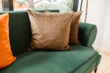 Pencerenin yanında yumuşak yastıklı yeşil kanepenin köşesi. Sessiz konfor, ev sıcaklığı ve günlük rahatlama tekstil dokusu ve doğal iç ışık ile yaratıldı.