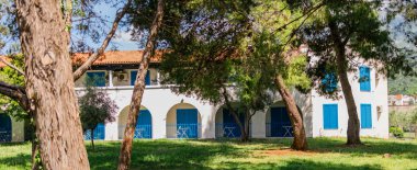 Bahçeyle çevrili mavi panjurları ve kemerleri olan beyaz bir bina. Akdeniz mimarisi, huzurlu tatil atmosferi ve otel terası.