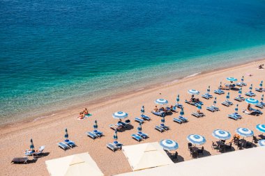 Adriyatik Denizi kıyısında şemsiyeli ve şezlonglu organize çakıl taşı plajı. Karadağ 'ın Budva kentinde yaz turizmi, kıyı konforu, tatil rutini ve deniz kenarındaki rahat yaşam tarzı.