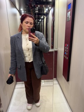 Asansörde aynalı selfie çeken bir kadın işaretli ceket ve kahverengi pantolon giyiyor. Sıradan bir anda günlük özgüven, kendini ifade etme ve modern şehir yaşam tarzı