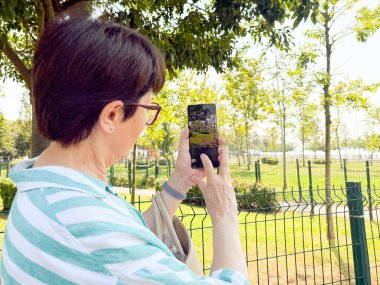 Elinde akıllı bir telefonla dışarıda fotoğraf çeken bir kadın. Açık bir park ortamında günlük yaşam, şehir eğlencesi ve kişisel bakış açısı