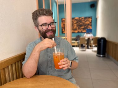 Ahşap kafe masasında buzlu meyve çayının keyfini çıkaran bir adam. Rahatlamış ruh hali, günlük rutin, ve modern içecek yaşam tarzı ile günlük ferahlatıcı.