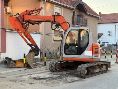 Gün batımında banliyödeki evlerin yanındaki iskan caddesinde çalışan kazıcı. Yol onarımı, kentsel altyapı ve inşaat faaliyetleri ve belediye hizmeti yoluyla mahalle gelişimi.