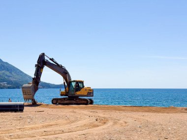Deniz ve dağların yakınındaki kumlu sahilde duran sarı bir kazıcı. Sahil şeridinde endüstriyel varlık, inşaat ve insan faaliyetleri