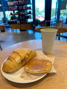 Sıcak kahvaltı sahnesi, kruvasan, krep ve ahşap bir masada servis edilen kibrit latte. Kafenin bulanık arka planı derinlik katar ve sıradan bir yemek ortamında sıcaklık ve rahatlama hissi uyandırır. Kahve dükkanı konsepti.