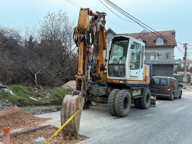 Evleri ve elektrik hatları olan bir banliyö yerleşim bölgesinde yol onarımı üzerinde çalışan kazıcı. Kentsel altyapı bakımı, belediye inşaat faaliyetleri, inşaat mühendisliği ve kamu hizmetlerinde iyileştirme