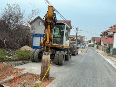 Evleri ve elektrik hatları olan bir banliyö yerleşim bölgesinde yol onarımı üzerinde çalışan kazıcı. Kentsel altyapı bakımı, belediye inşaat faaliyetleri, inşaat mühendisliği ve kamu hizmetlerinde iyileştirme