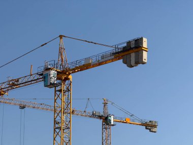 Gökyüzünün altında bir inşaat alanının üzerinde yükselen kule vinçleri. Mühendislik, geliştirme, kentsel genişleme, endüstriyel ilerleme, yapısal planlama ve modern yapı faaliyetlerinin geometrisi