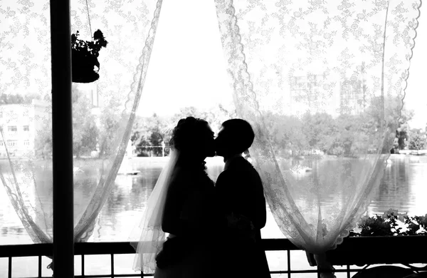 Joyeux jeunes mariés s'embrassant à l'intérieur. Brunette mariée et marié debout près des silhouettes de fenêtre de heureux — Photo