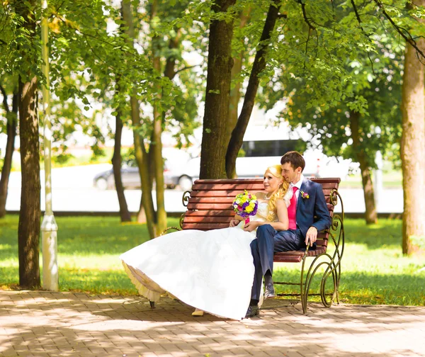 公園のベンチに座っているエレガントな新郎新婦白ドレスでスタイリッシュな結婚式カップル金髪花嫁. — ストック写真
