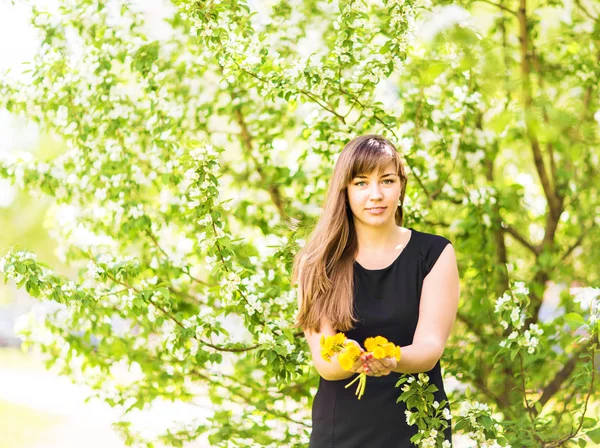 Porträt eines schönen Mädchens mit einem Strauß gelber Löwenzahne im Freien im Frühling, Fokus auf Augen, Apfelblütenhintergrund — Stockfoto