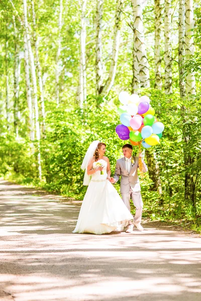 Beautiful bride holding bunch of balloons in the park. Couple of bride and groom with balloons. Newlyweds with balloons outdoors — Stock Photo, Image