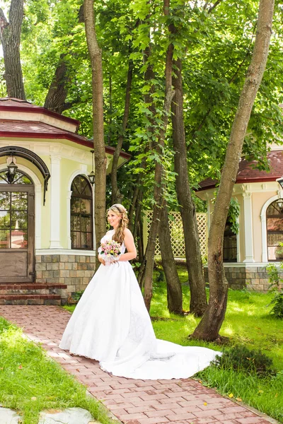 Krásu nevěsty ve svatební šaty s kytice a krajkový závoj na povaze. Krásný model dívka v bílých svatebních šatech. Ženský portrét v parku. Žena s účes. Roztomilá dáma venku — Stock fotografie