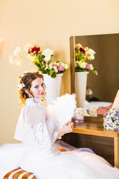 Bela noiva em vestido de noiva com ventilador sentado em seu quarto perto do espelho — Fotografia de Stock