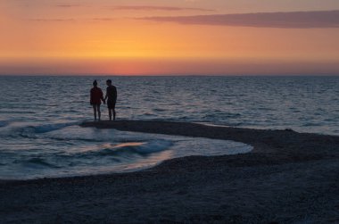 Bir adam ve bir kadın deniz kıyısında el ele tutuşuyorlar. Küçük dalgalar. Pembe günbatımına karşı karanlık siluetler. Romantik akşam. Yaz zamanı. Aşk hikayesi.