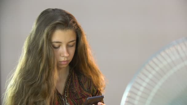 Όμορφο κορίτσι που κάθεται σε μια καρέκλα κοντά το ανεμιστήρα και γράφει sms στο κινητό — Αρχείο Βίντεο