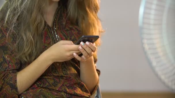 Bella ragazza seduta su una sedia vicino al ventilatore e scrive sms sul cellulare — Video Stock