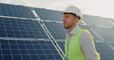 Üniformalı yakışıklı erkek mühendisin yürümesi ve güneş enerjisi santraline bakması. Sert miğferli bir adam taslak taşırken nesneleri inceliyor. Yeşil enerji kavramı.