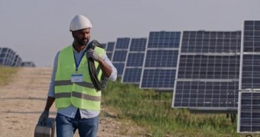 Erkek sakallı mühendisin kabil ve çanta taşıdığı ekin manzarası. Güneş panelleri çiftliğinde yürüyen kasklı ve üniformalı melez bir yarış adamı. Yenilenebilir, yeşil enerji ve yenilikler kavramı.