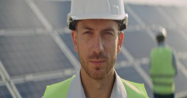 Kafasını çevirip kameraya bakan kasklı erkek mühendisin portresi. Üniformalı neşeli adam güneş enerjisi santralinde dururken gülümsüyor. Güneş istasyonu gelişimi kavramı. Anamorfik lens.