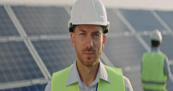 Mužský inženýr v ochranné přilbě otáčí hlavu a dívá se do kamery. Muž v uniformě se usmívá, když stojí ve sluneční elektrárně. Koncept rozvoje solárních stanic. Anamorfní objektiv. — Stock video