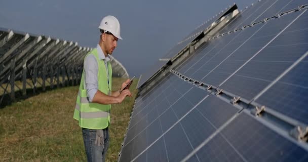 Mužský inženýr zadává data při použití tabletu a dotýká se solárního panelu. Muž v uniformě kontroluje práci fotovoltaických článků a usmívá se, zatímco stojí na zelené energetické farmě. Anamorfní objektiv. — Stock video