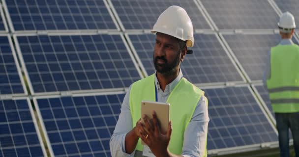 Trabalhador do sexo masculino olhando para tela tablet e do que à parte. Homem de capacete duro e uniforme em pé na fazenda de energia renovável e usando gadget. Painéis solares no fundo. Lente anamórfica. — Vídeo de Stock