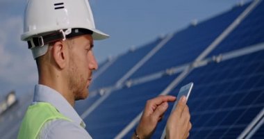 Erkek işçi tablet ekranına ve güneş paneline bakıyor. Sert kasklı ve üniformalı bir adam yenilenebilir enerji çiftliğinde duruyor ve alet kullanıyor. Yenilik kavramı. Anamorfik lens.