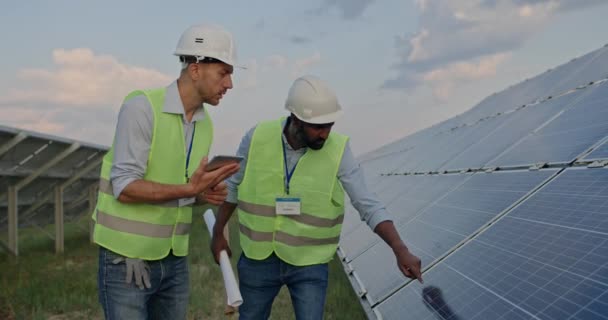 Ingeniero masculino comprobando la operación de paneles solares fotovoltaicos y hablando con su colega. Hombre en casco duro y uniforme usando tableta para introducir datos. Concepto de energía renovable. — Vídeo de stock