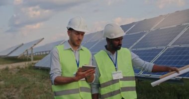 Erkek mühendis tomarı tablet kullanan iş arkadaşına emir veriyor. Üniformalı ve kasklı iki adam güneş çiftliğinde yürürken projeyi tartışıyorlar. Yeşil enerji kavramı.