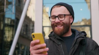 30 'lu yaşlarda sakallı bir adamın telefon ekranına bakıp sokakta yürürken gülüşünü yakından izleyin. Akıllı telefon kullanırken sosyal medya haberlerini kaydıran gözlüklü yakışıklı adam.